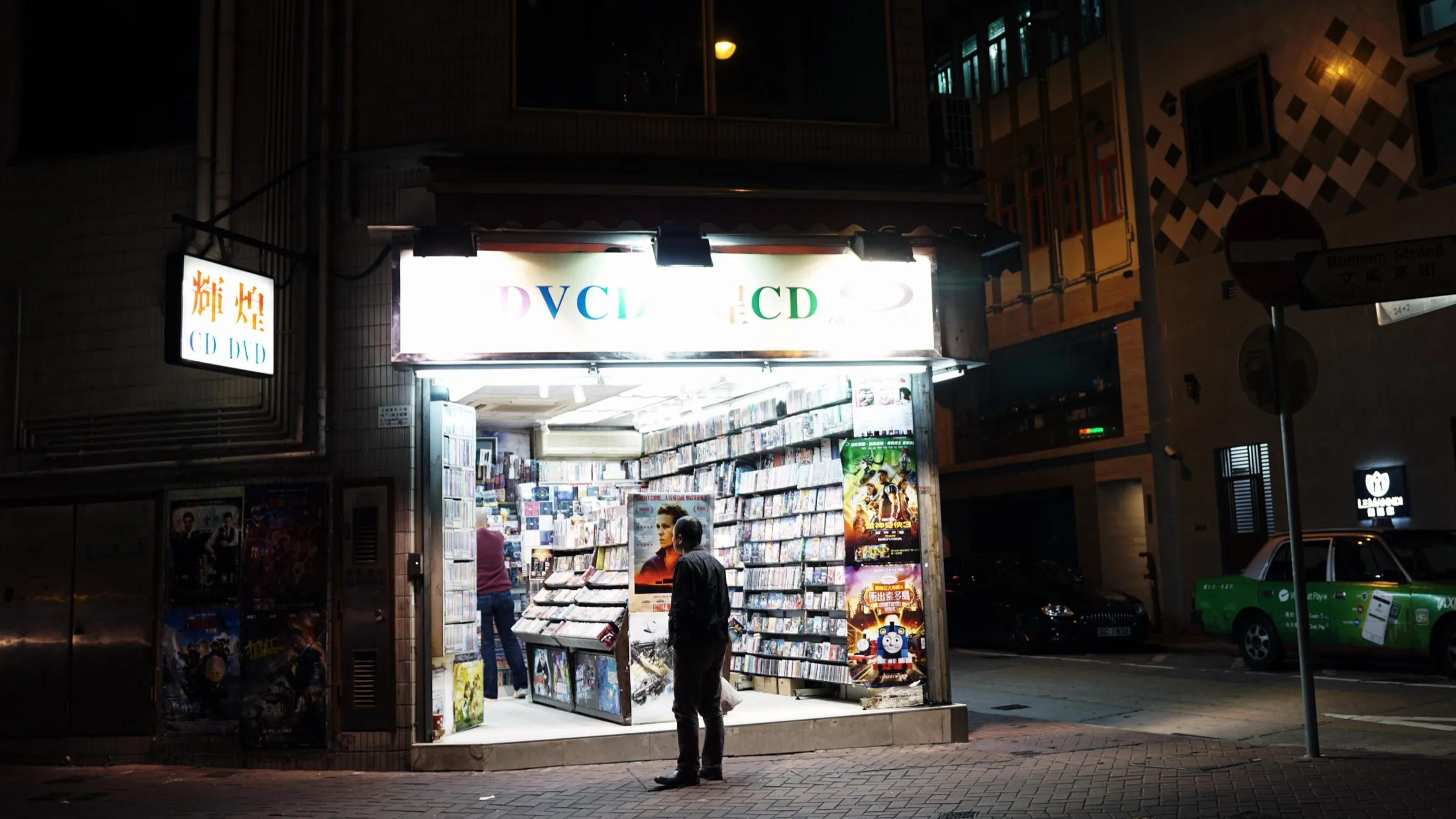 DVD와 CD를 파는 가게가 밤에 불을 밝히고, 한 남자가 밖에 서서 안을 들여다본다.